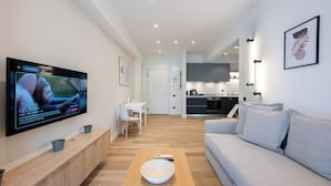 A modern living room with a flat-screen TV, a wooden coffee table, a grey sofa, and a kitchen in the background.