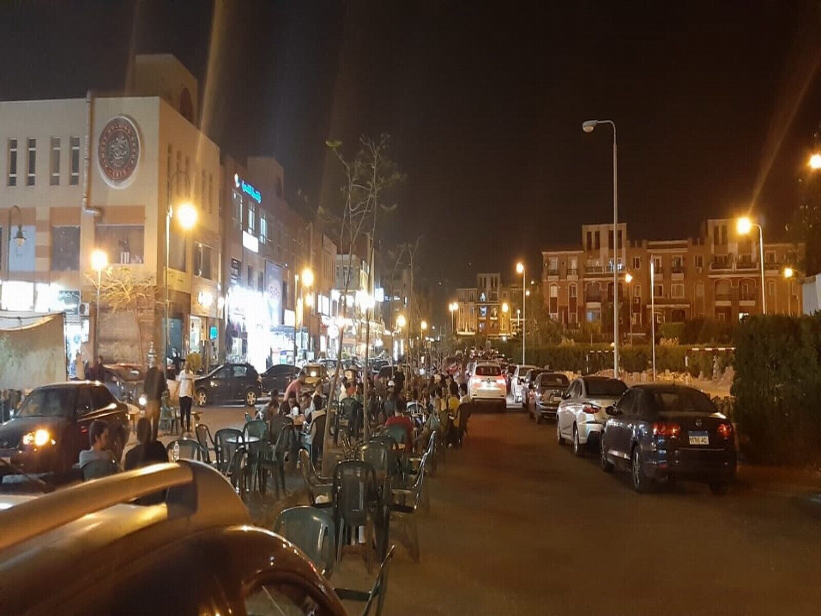 Eine Straße mit geparkten Autos und Außenbestuhlung, was auf einen Gastronomiebereich oder ein Café hindeutet.