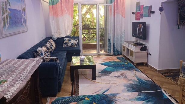 A modern living room with a sofa, a coffee table, and a TV. There is a sliding glass door leading to a balcony with a view of greenery. - Cozy apartment close to the Giza Pyramids' (El Sheikh Zayed City)