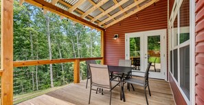 Eine Holzterrasse mit Tisch und Stühlen, eine Tür, die zu einer grünen Terrasse führt, und Blick auf einen Wald.
