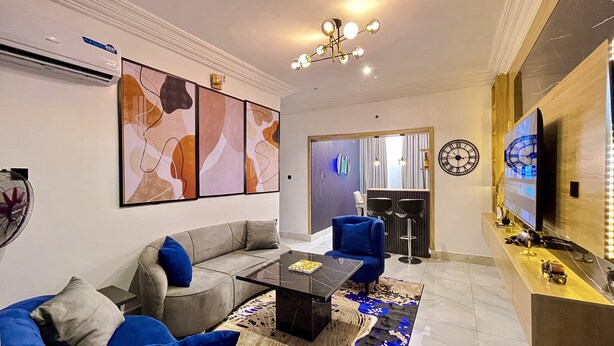A modern living room with a sofa, a coffee table, and a blue armchair. There is a TV on the right and a clock on the wall. - Luxury 3BR Ensuite Apartment 24/7 Power Fast WiFi Secure Estate Ikeja Lagos (Ikeja)