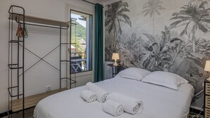Ein Hotelzimmer mit Bett, Fenster mit Blick auf Grünflächen, Wand mit tropischem Wandbild und ein Metallregal mit Handtüchern.