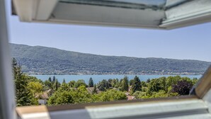 Blick aus einem Fenster auf einen See und Berge, mit Bäumen und Häusern im Vordergrund.