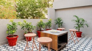 Un pequeño espacio al aire libre con mesa de madera y cuatro taburetes, rodeado de plantas en macetas y suelo de baldosas.