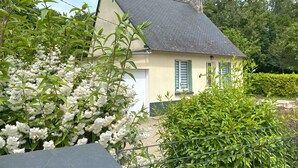 Una casa pequeña con techo inclinado, rodeada de exuberante vegetación y plantas en flor.