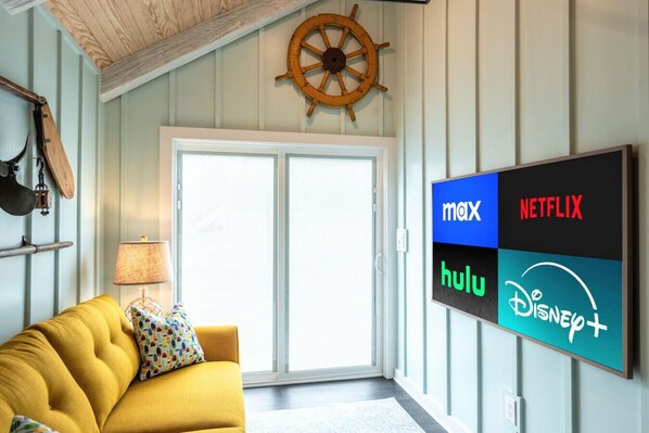 Ein Wohnzimmer mit gelbem Sofa, Holzboden, einem großen Fenster und einem an der Wand befestigten Fernseher, der Logos von Streaming-Diensten anzeigt.