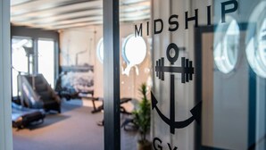 Une salle de sport avec un logo sur la porte en verre, des appareils d’exercice et une plante.