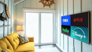 Un salon avec un canapé jaune, un téléviseur fixé au mur qui diffuse des services de streaming, et un volant de navire en bois au-dessus d’une porte coulissante en verre.