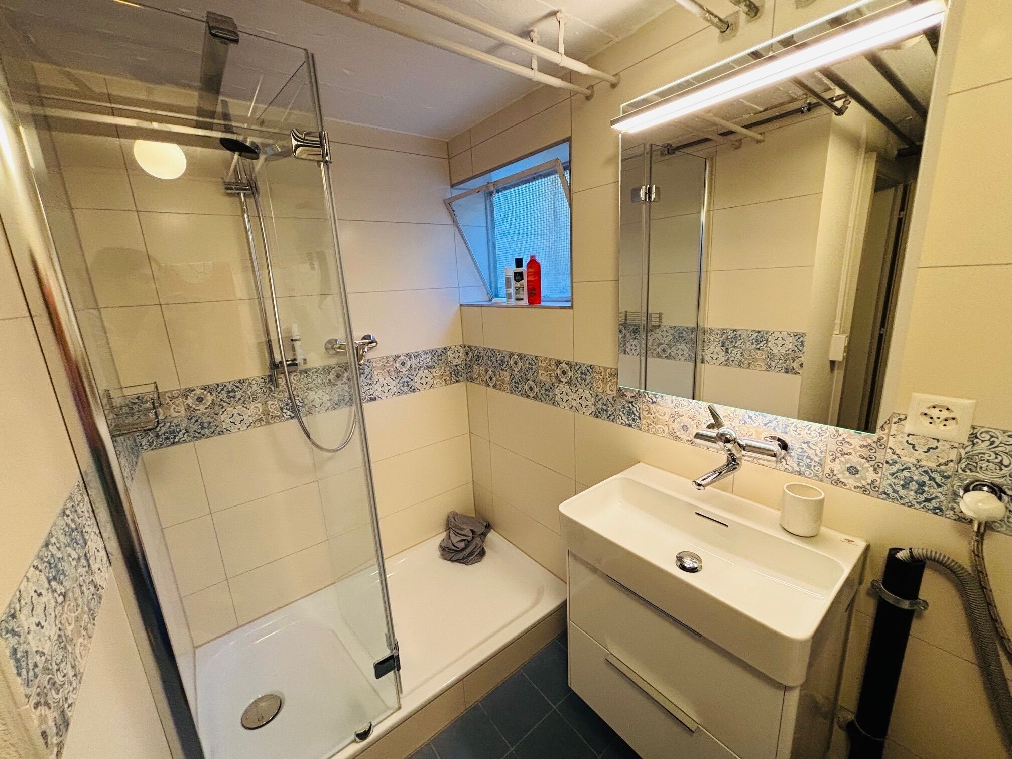 A bathroom with a glass-enclosed shower, a bathtub, a sink with a mirror, and a window with blinds.