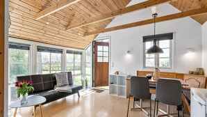 Un vaste espace de vie doté d’un plafond en bois, d’un canapé noir, d’une table à manger avec des chaises et de grandes fenêtres offrant une vue sur l’extérieur.