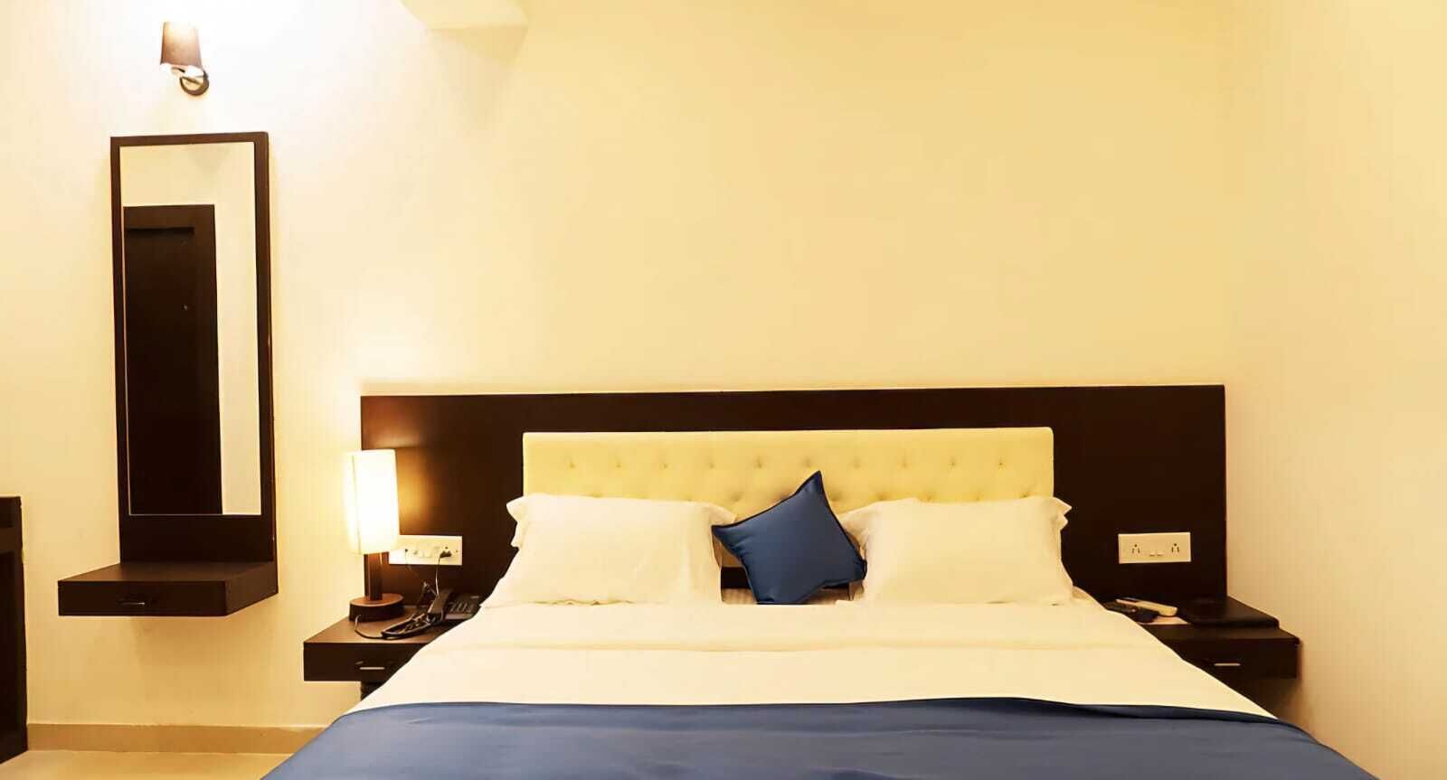 A hotel room with a large bed, a nightstand with a lamp, a mirror, and a wall-mounted light fixture.