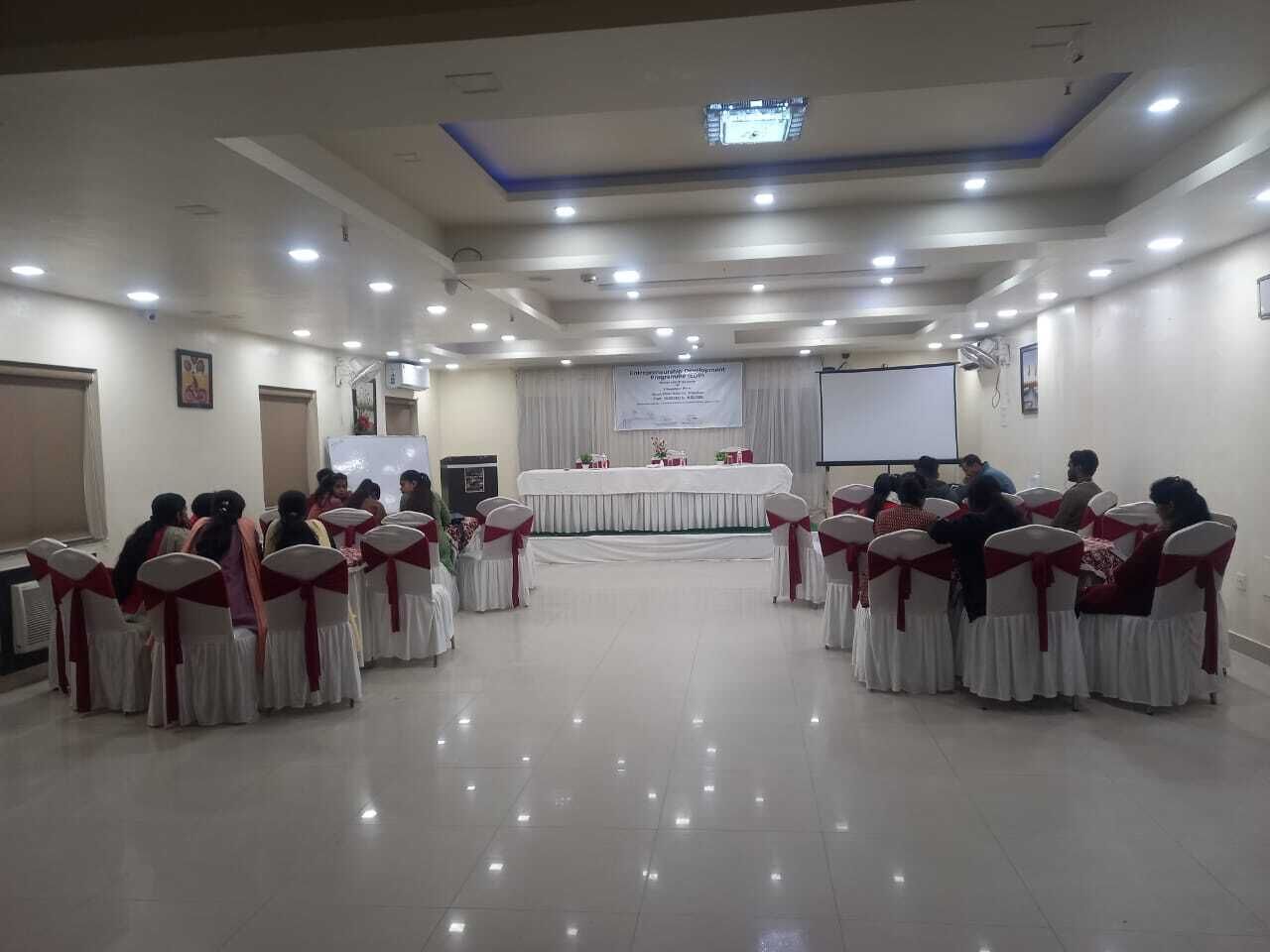 Un salón de conferencias con mesas y sillas redondas, un escenario con un atril y una pantalla de proyector.