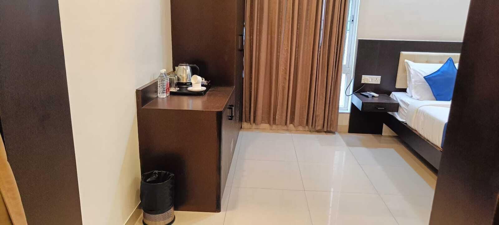 A hotel room with a bed, a nightstand, a desk with a kettle, a trash bin, and a window with curtains.