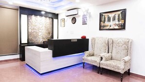 A reception area with a white reception desk, a black counter, a floral armchair, and a framed cityscape painting on the wall. - Hotel Max INN (Bhagalpur)