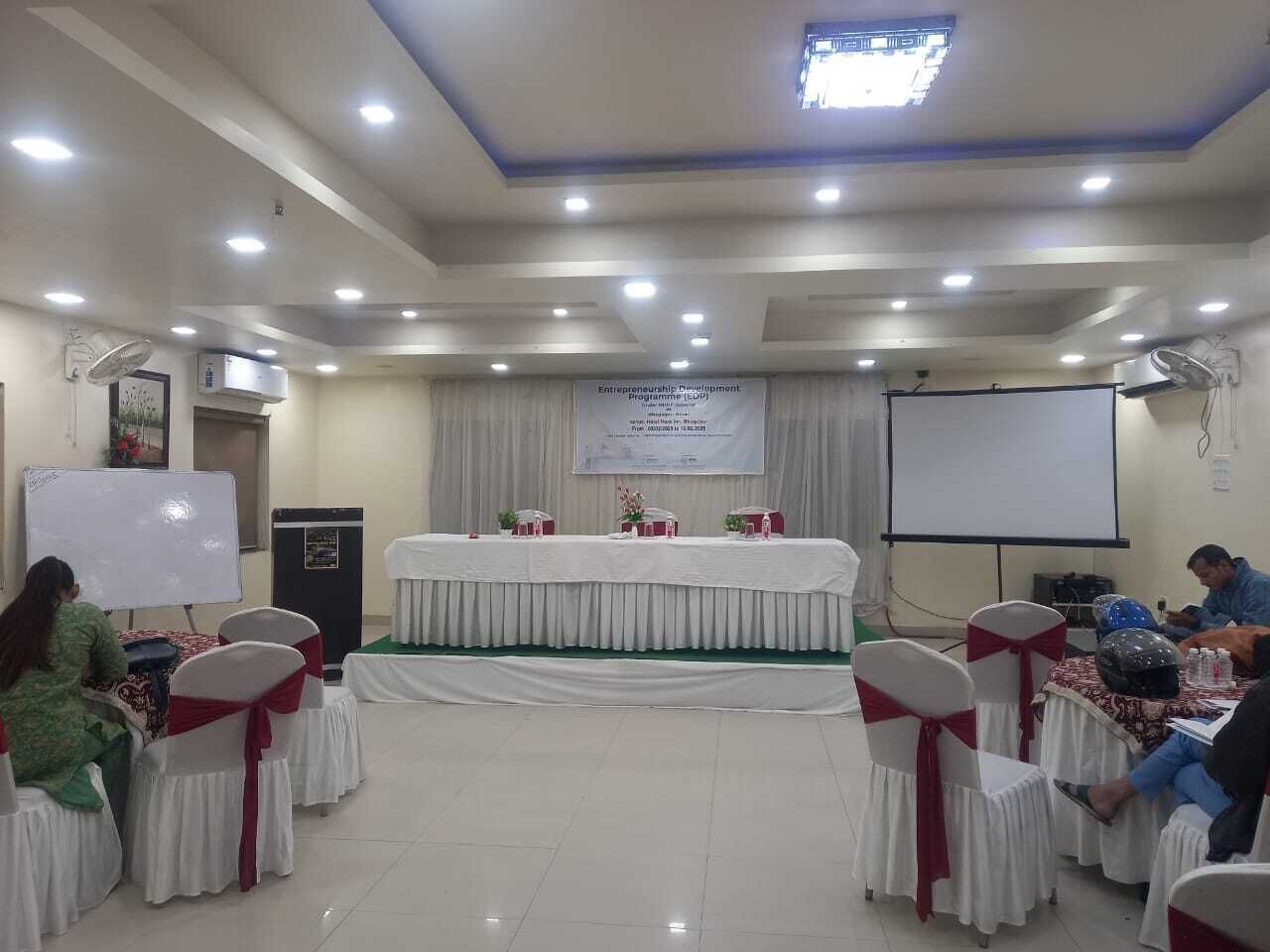 Un salón de conferencias con un escenario, sillas blancas con cintas rojas, una pizarra y un cartel.