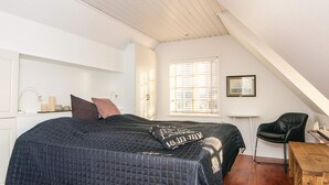 Une chambre à coucher avec un grand lit, un bureau en bois, une chaise, une fenêtre avec des stores et un tableau encadré au mur.