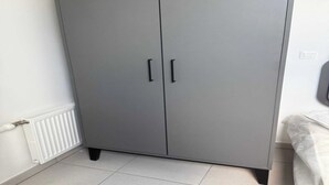 Un armario gris con dos puertas y tiradores negros, situado junto a un radiador blanco y una cama con cubrecama blanco.