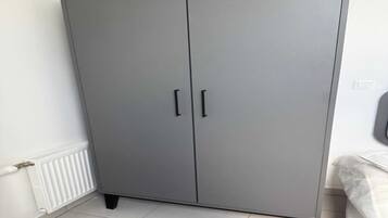 A grey wardrobe with two doors and black handles, positioned next to a white radiator and a bed with a white cover.