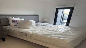Habitación de hotel con una cama cubierta en plástico, cabecera gris, mesita de noche con lámpara y vista al exterior por una puerta.