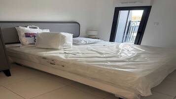 A hotel room with a bed covered in plastic, a grey headboard, a bedside table with a lamp, and a view of the outside through a door.
