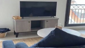 Una sala de estar con una televisión de pantalla plana sobre un mueble bajo, un sofá con cojines oscuros, una pequeña mesa redonda y vistas a un balcón con barandilla.