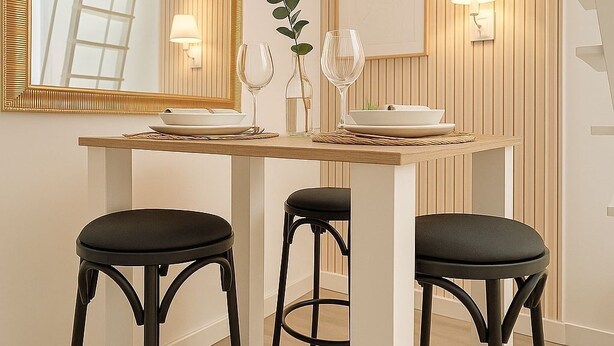 A dining area with a wooden table, two black stools, a mirror with a gold frame, and wall-mounted lamps. - Charming apartment for seasonal rental (Madrid)
