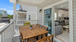 Una terraza de madera con una mesa y sillas, una puerta corrediza de vidrio que da a una sala de estar con sofá y mesa de centro, y vistas del horizonte de la ciudad.