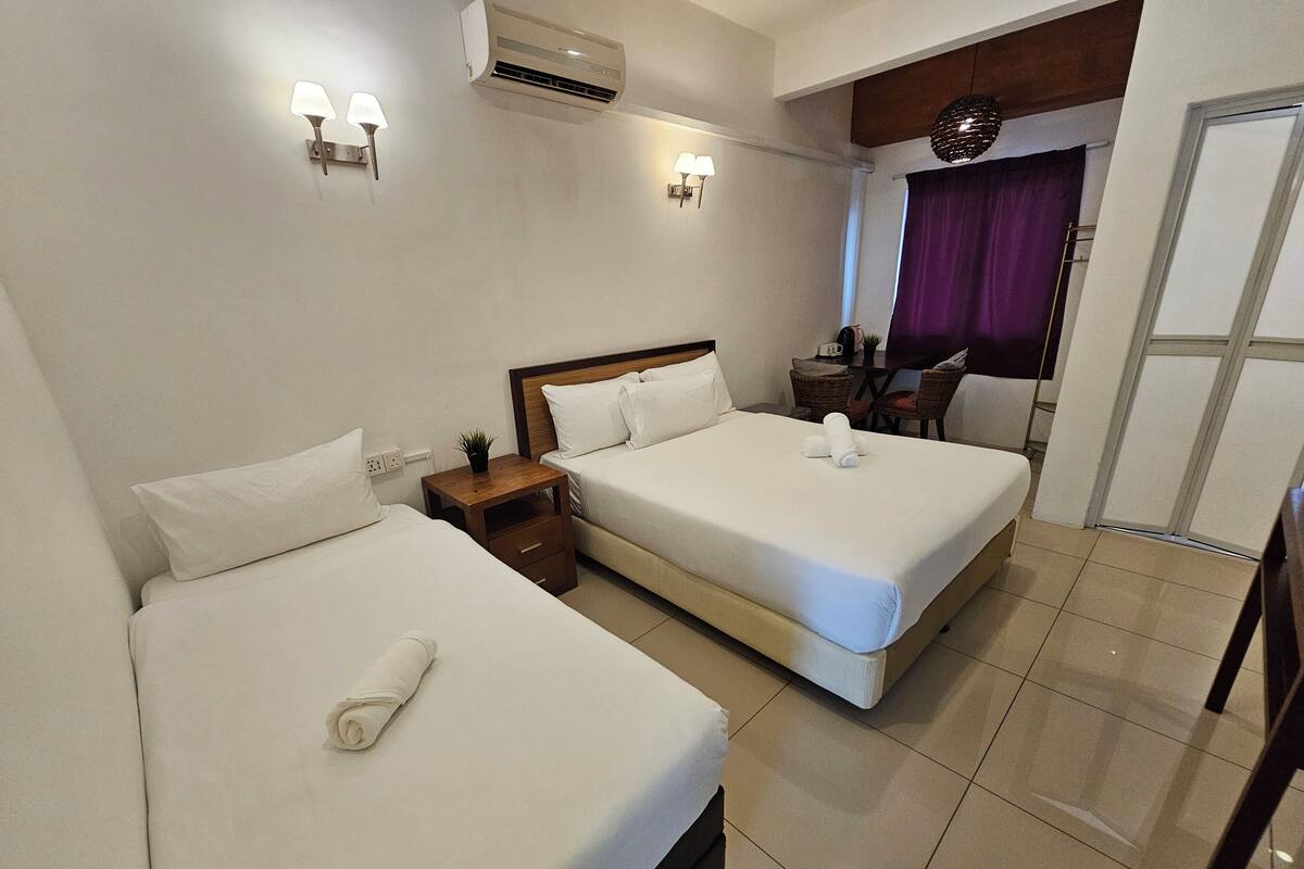 A hotel room with two beds, a small table with a plant, a desk with a chair, and a window with purple curtains.