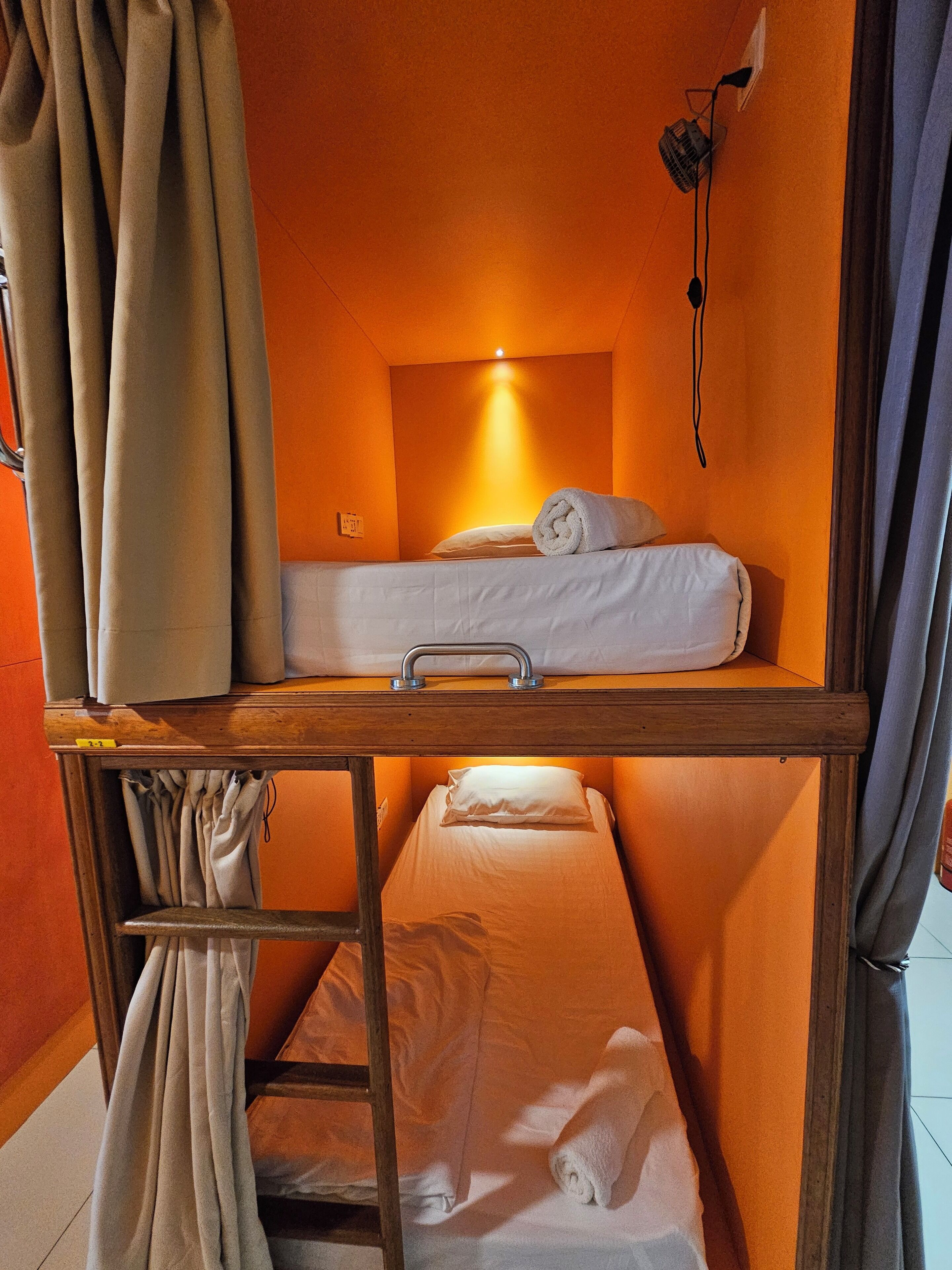 A bunk bed room with a wooden frame, a single bed on top, and a lower bunk. There is a small shelf with a lamp and a power outlet on the wall.