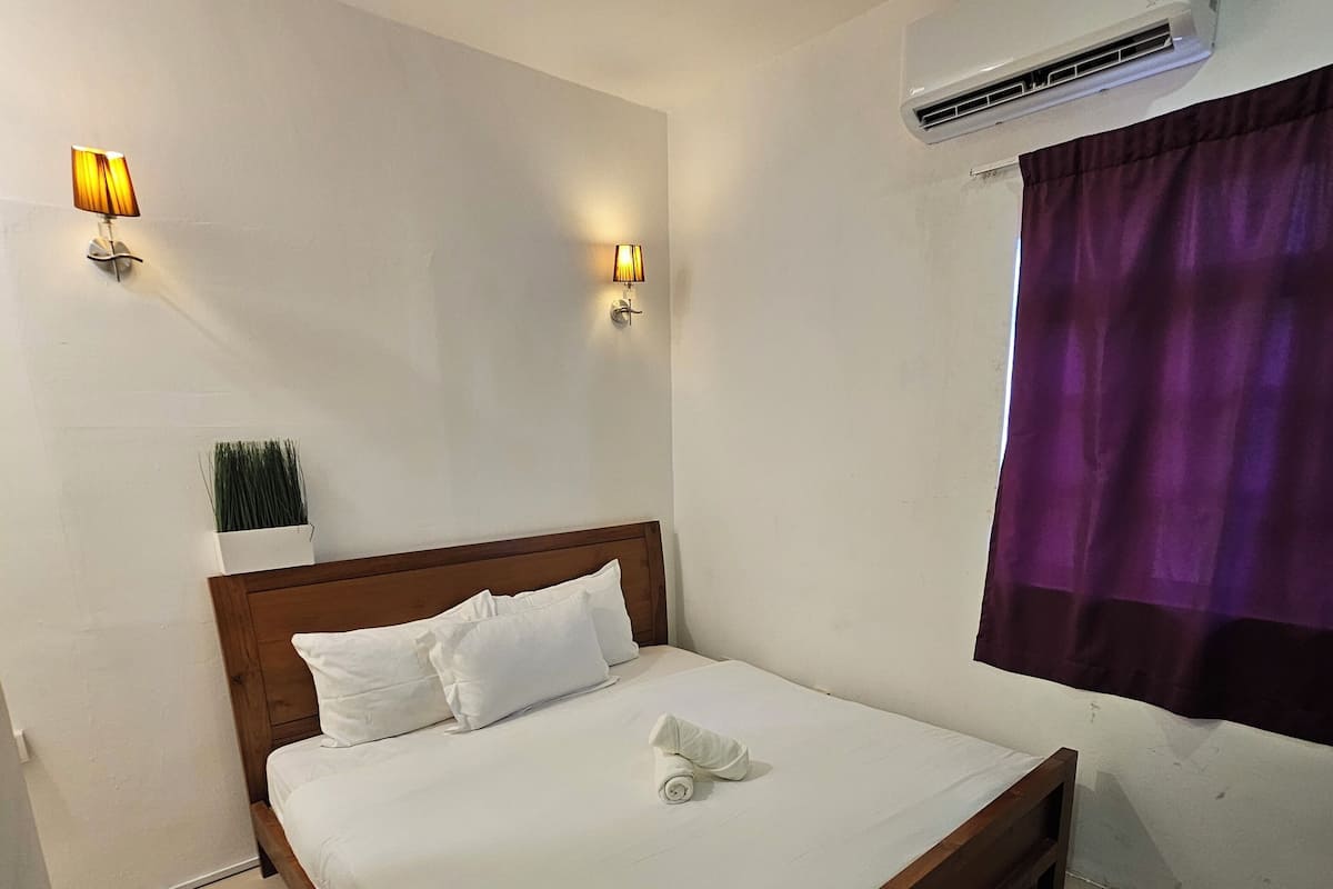 A neatly made bed with white linens, a wooden headboard, two wall-mounted lamps, and an air conditioning unit.