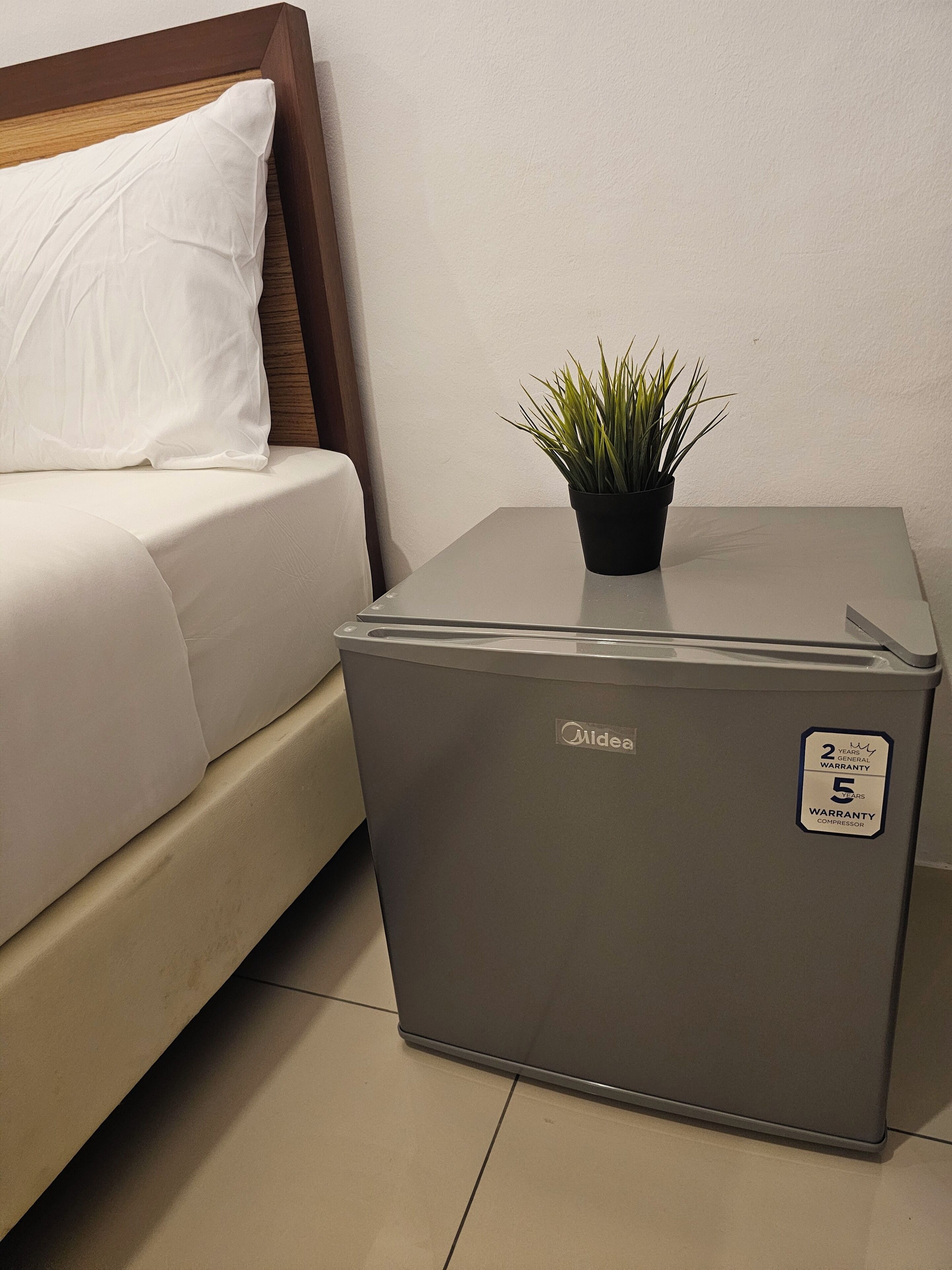 A compact refrigerator with a two-year general warranty and a five-year compressor warranty sticker, placed on a tiled floor next to a bed with a white pillow and a small potted plant.