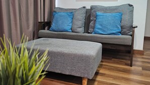 A modern living room with a grey sofa, blue cushions, a grey ottoman, and a wooden coffee table. A large abstract painting is on the wall, and a potted plant is in the foreground. - Heliconia by Treebridge (George Town)
