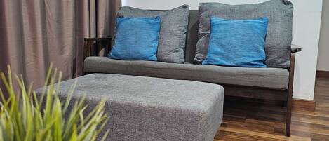 A modern living room with a grey sofa, blue cushions, a grey ottoman, and a wooden coffee table. A large abstract painting is on the wall, and a potted plant is in the foreground.