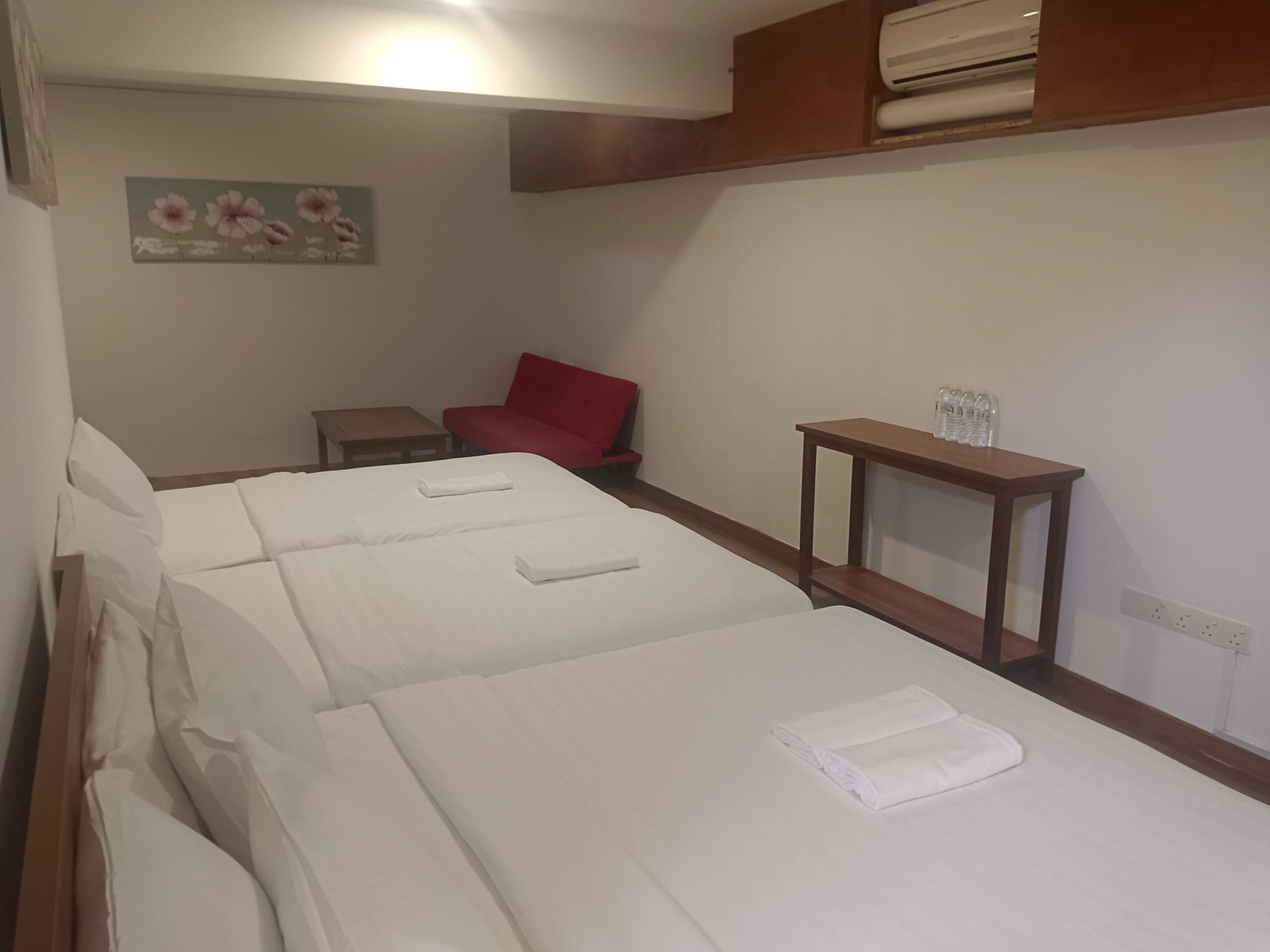 A hotel room with three beds, a red chair, a small table, and a wall-mounted air conditioner.