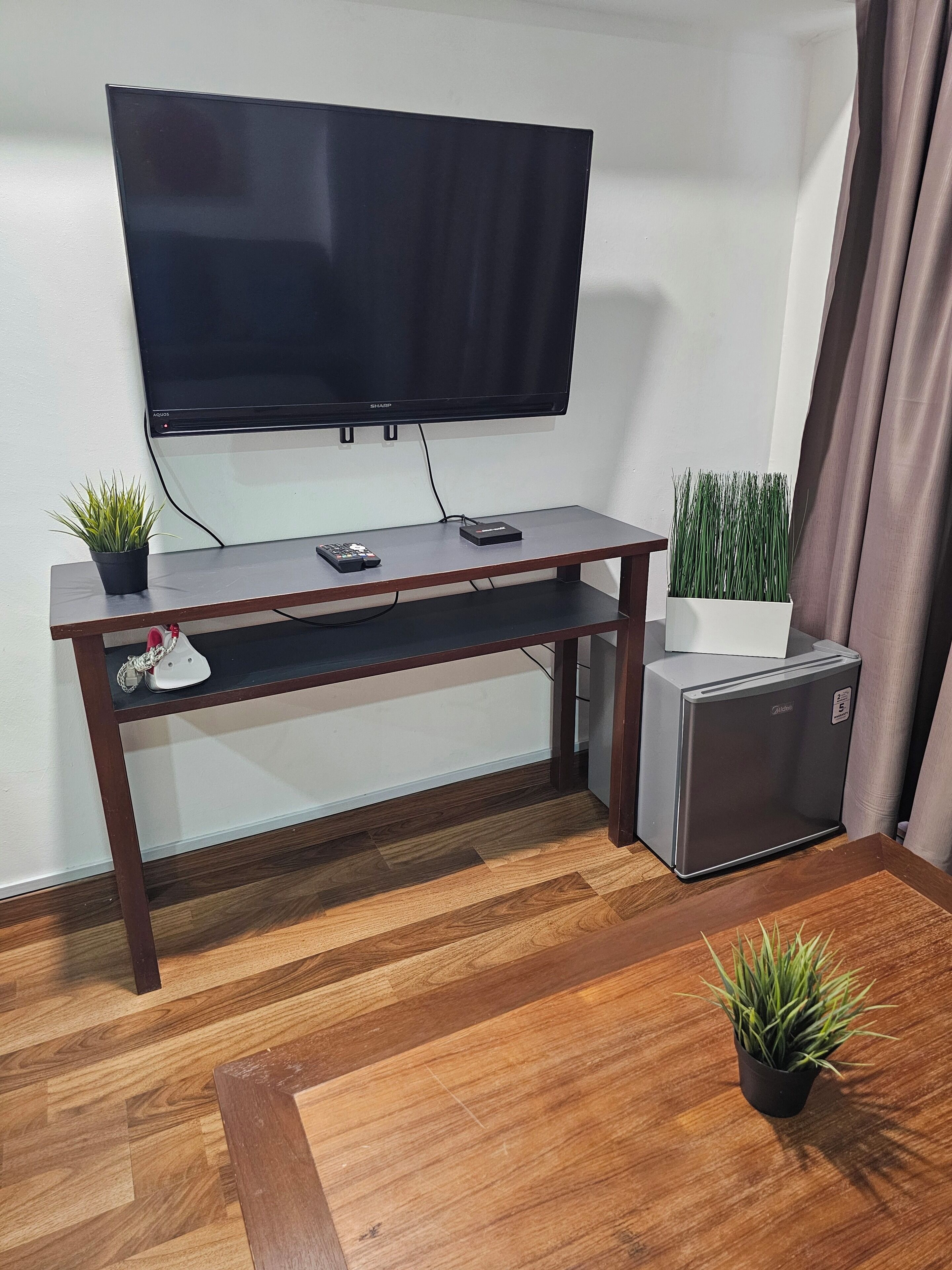 A room with a TV mounted on the wall, a small refrigerator, a wooden table, and two potted plants.