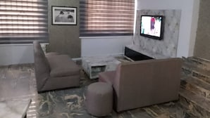 A living room with a sofa, ottoman, and a flat-screen TV mounted on a marble wall. - Morada Hotels (Lagos)