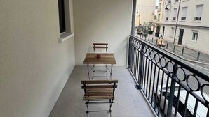Un balcon avec une table en bois et deux bancs, une fenêtre, et une vue sur une rue bordée d’immeubles et de voitures garées.