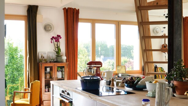 A modern kitchen with a large island, wooden beams, and a view of greenery outside.