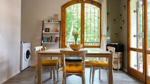 Un comedor con mesa y sillas de madera, una estantería, una lavadora y un amplio ventanal con vistas a zonas verdes.