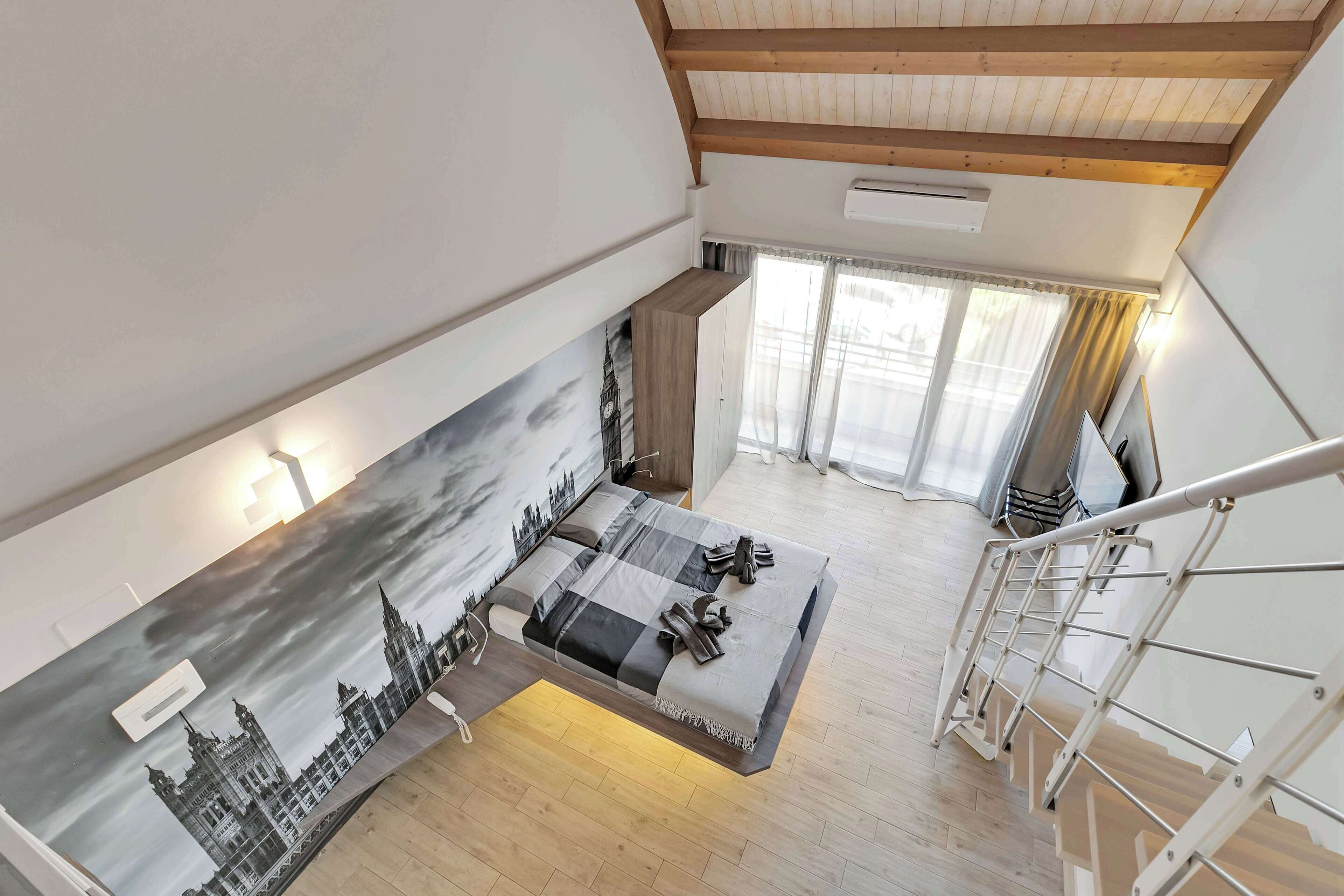 Une pièce spacieuse avec un plafond haut, un lit recouvert d’une couverture à carreaux, un tableau représentant un paysage urbain et un escalier avec une rampe en métal.