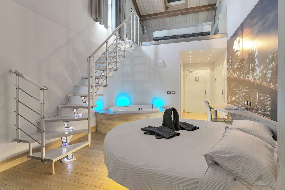 Une chambre d’hôtel moderne comprenant un lit, une salle de bain avec baignoire, un escalier et une fresque murale représentant un paysage urbain.