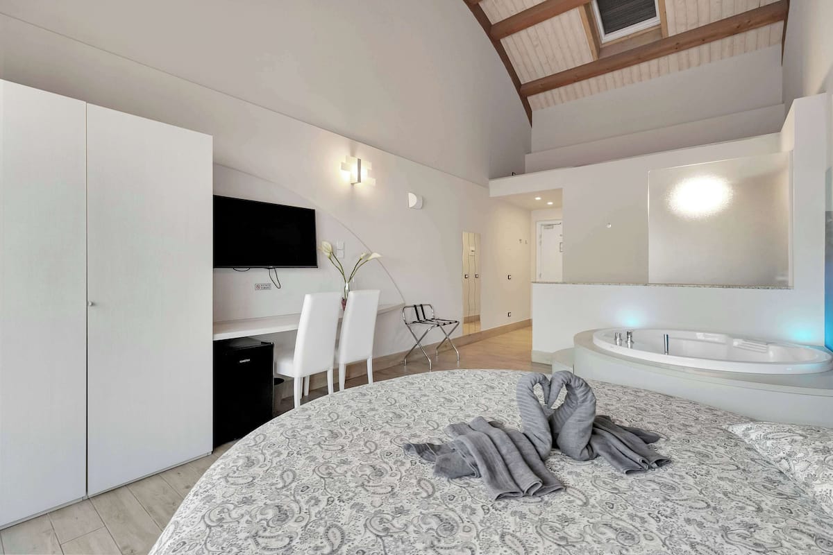 A modern bedroom with a large bed, a built-in desk, a flat-screen TV, and a separate bathroom with a bathtub.