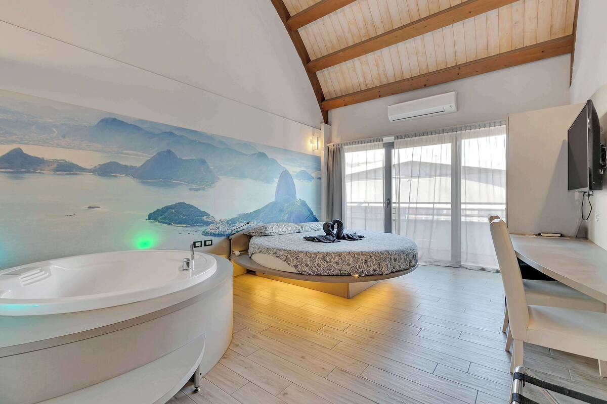 A modern bedroom with a large bed, a bathtub, and a scenic mural on the wall.