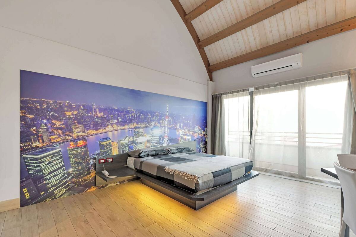 A modern bedroom with a cityscape mural, a bed with a headboard, and a large window with curtains.