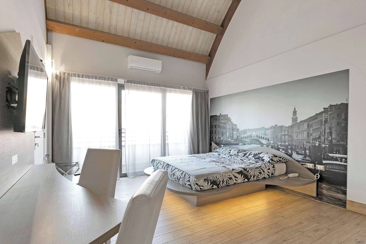 A modern bedroom with a bed, a dining table, and a chair. There is a large window with curtains, an air conditioning unit, and a wall-mounted TV.