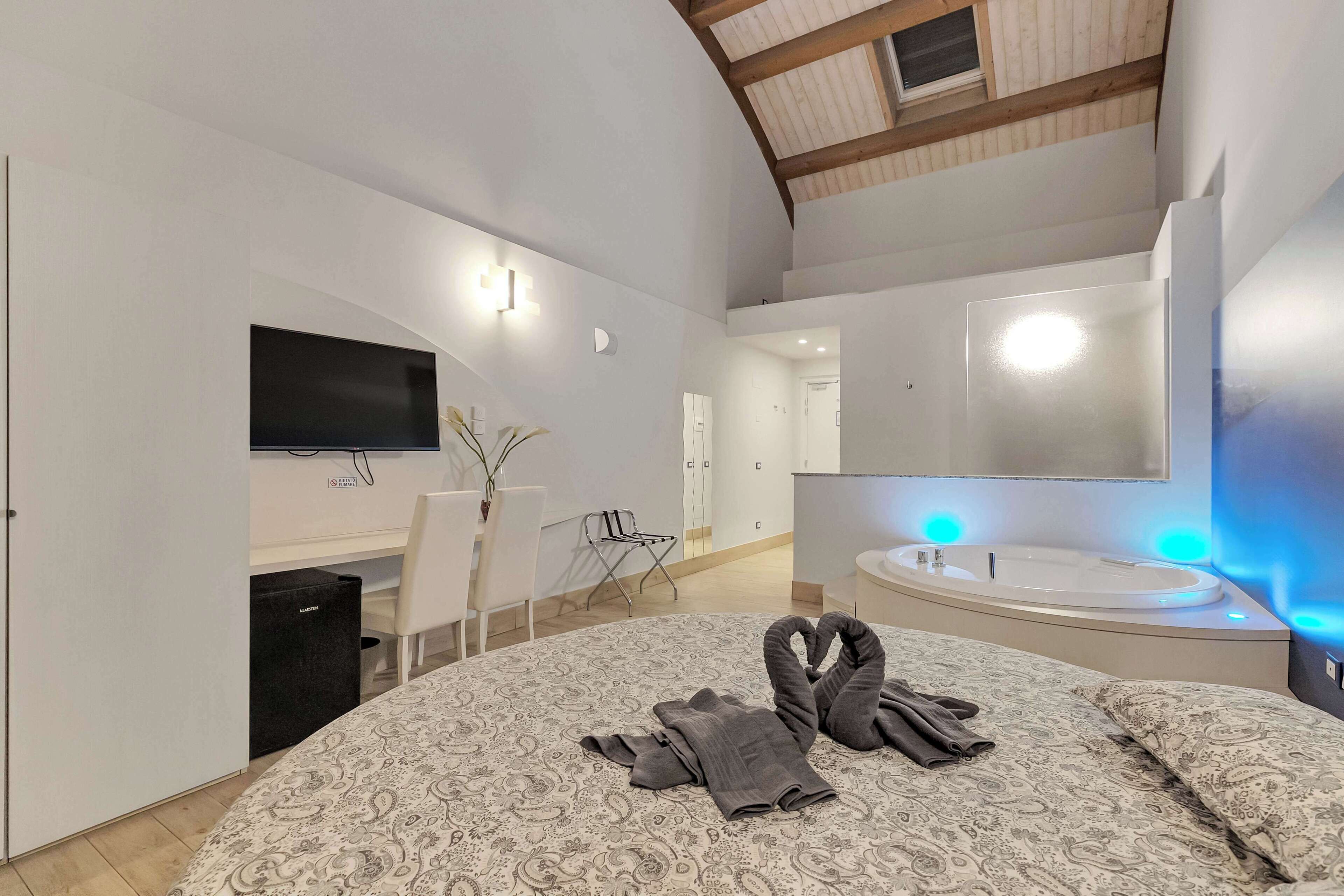 Habitación de hotel moderna con una cama grande, un escritorio con silla, un televisor de pantalla plana, un espejo amplio y una bañera con iluminación azul.
