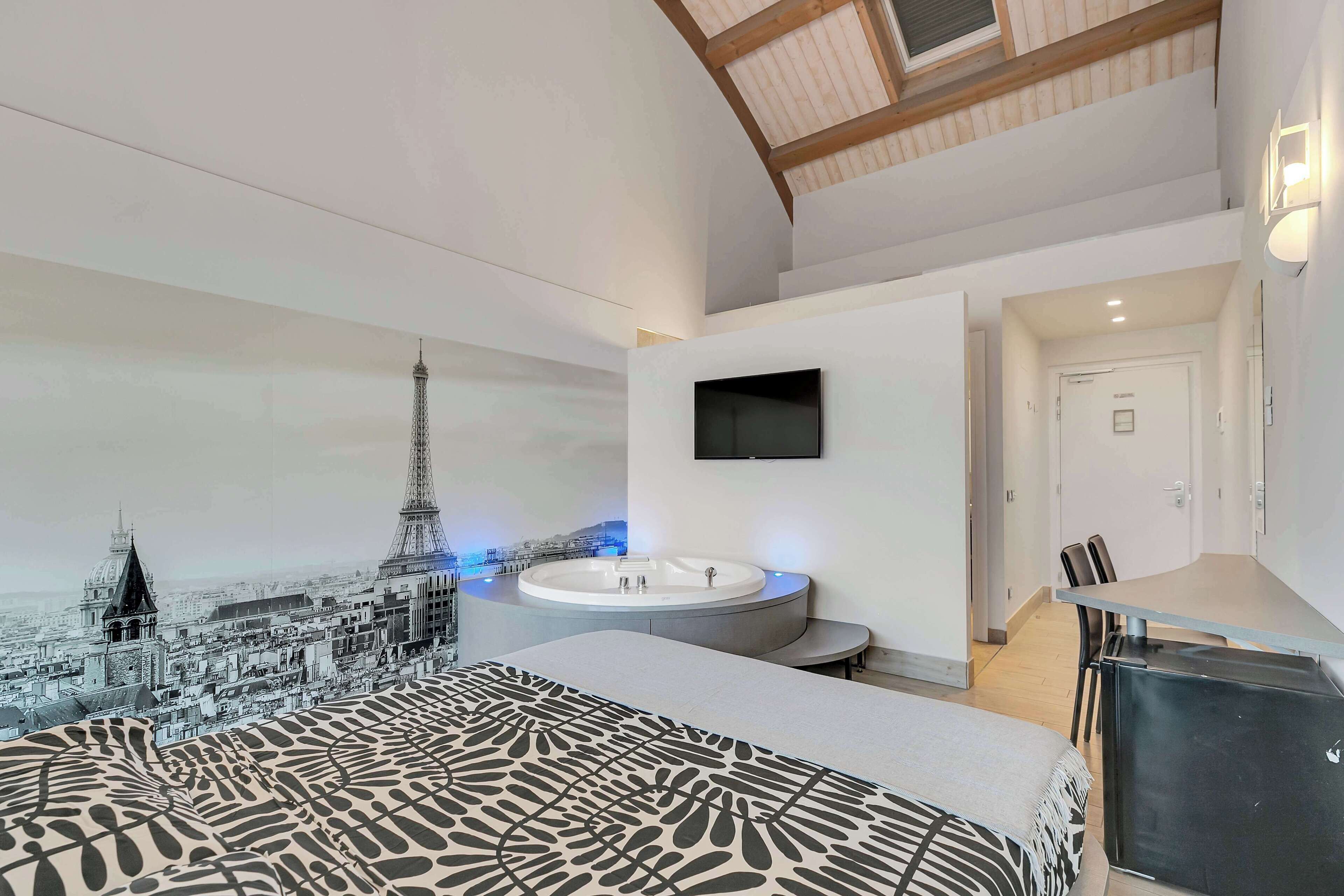 A bedroom with a bed, a large window, a wall-mounted TV, and a bathroom with a bathtub and sink.