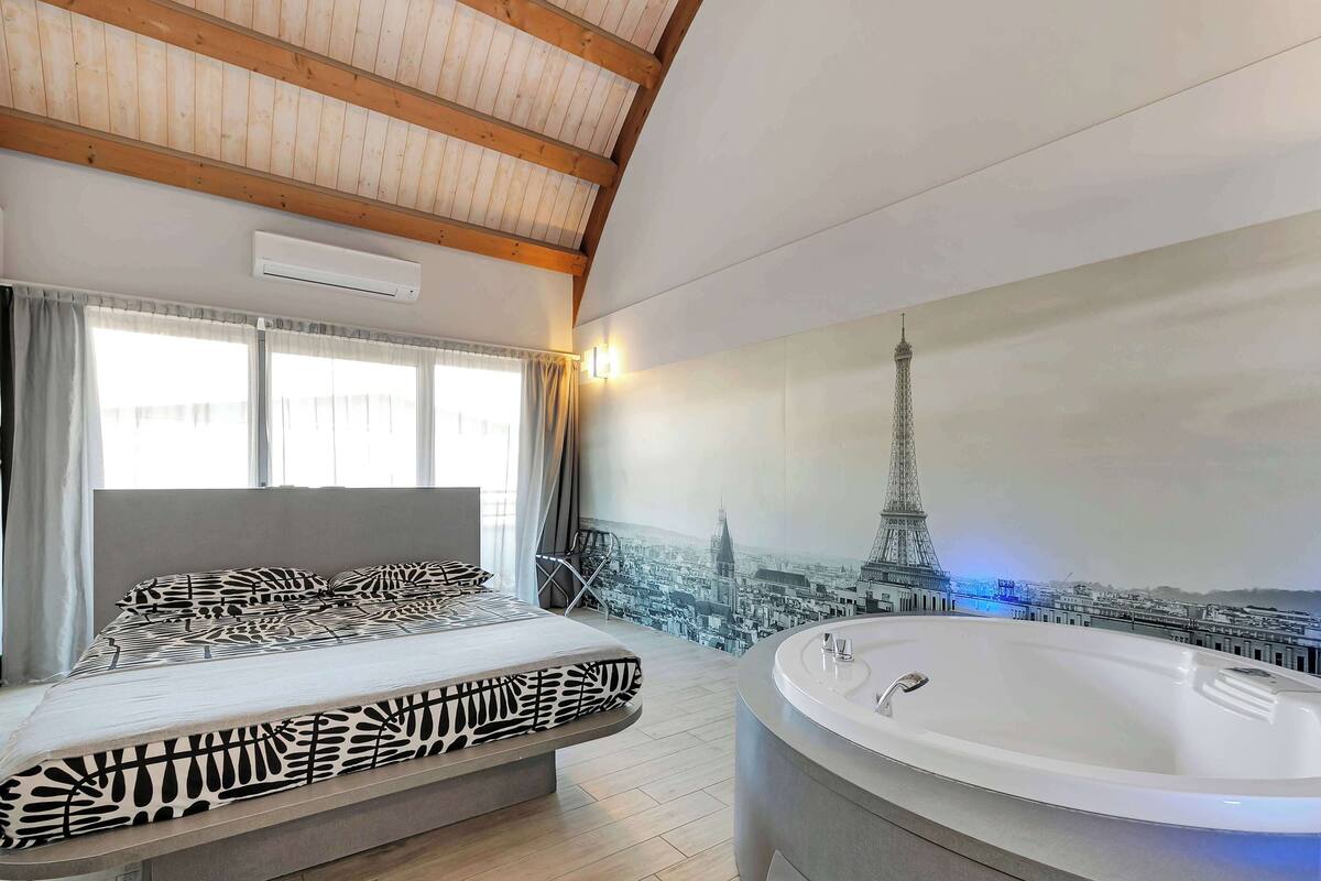 A bedroom with a bed, a large window, a wall-mounted air conditioner, and a bathtub.