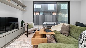 Ein modernes Wohnzimmer mit einem Flachbildfernseher, einem hölzernen Couchtisch, einem grünen Sofa und einer Glastür, die zu einem Außenbereich führt.