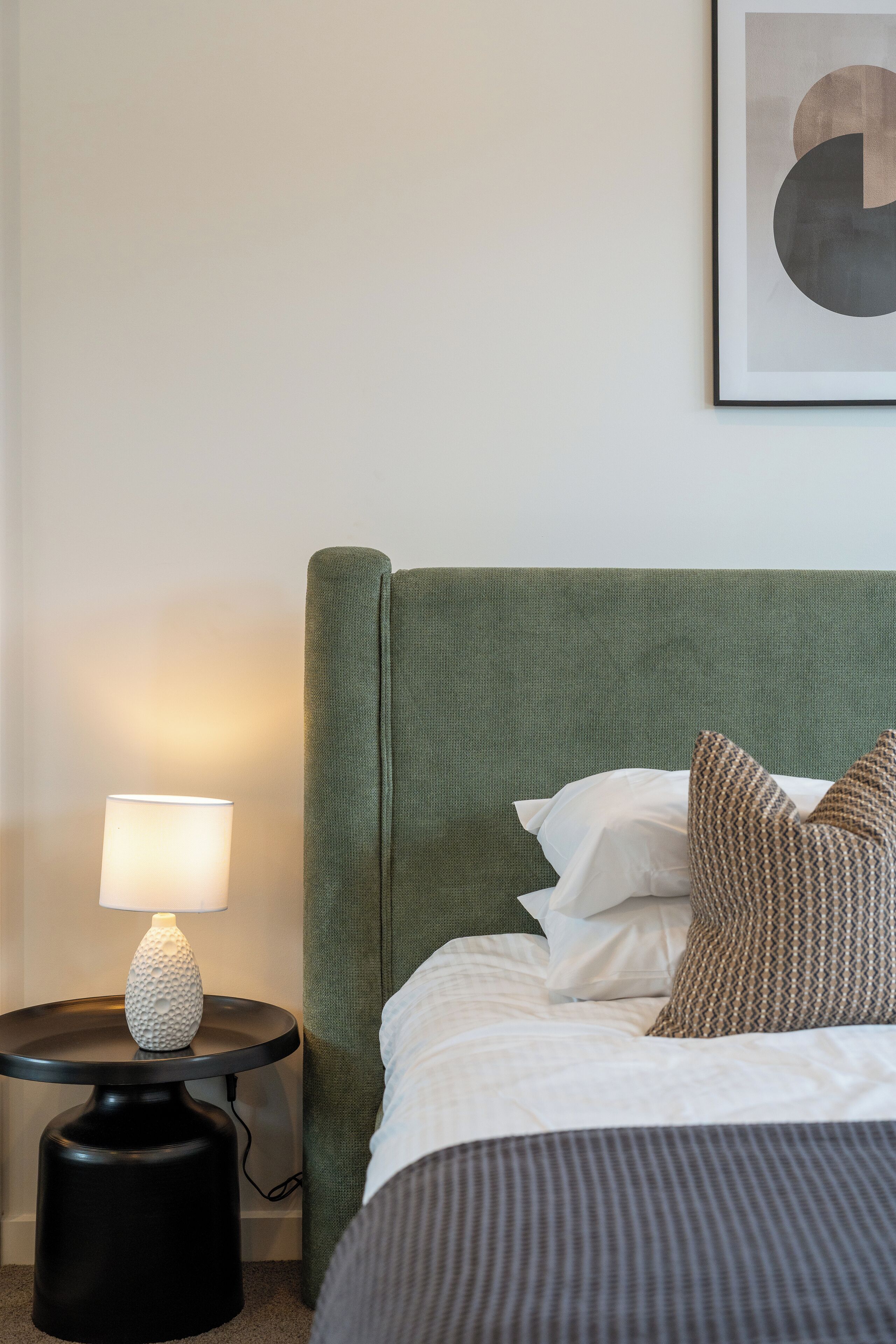 A neatly made bed with a green headboard, a bedside lamp, and a framed abstract artwork on the wall.